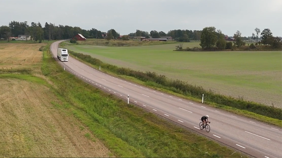 Drone-view over rural road where a long combination vehicle is approaching a bicyclist from behind.